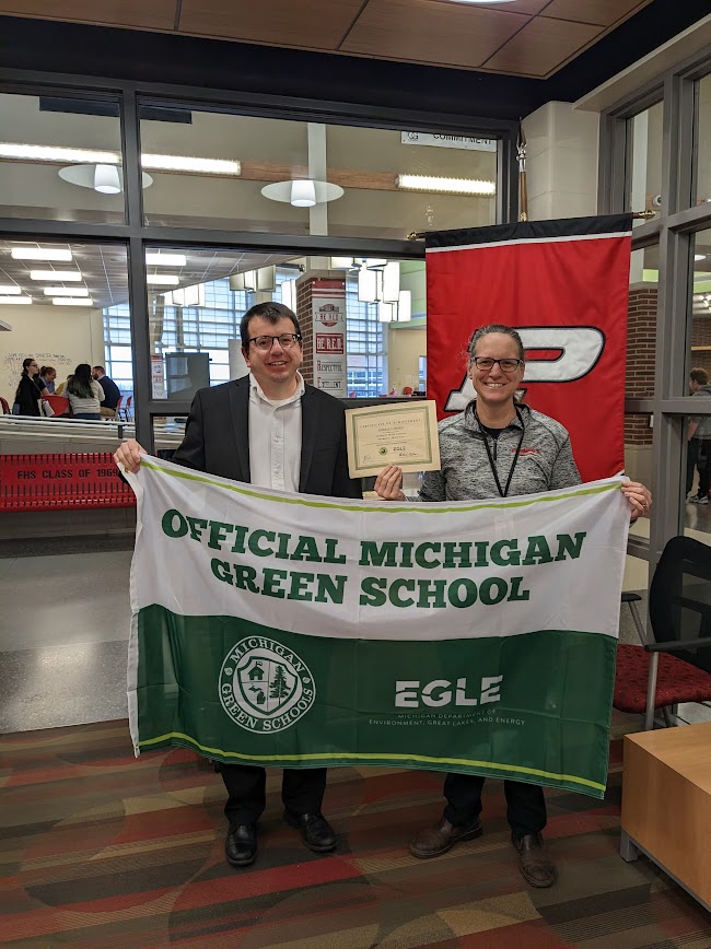Man and woman, stand in school office holding Green and white flag and Green School Certificate.
